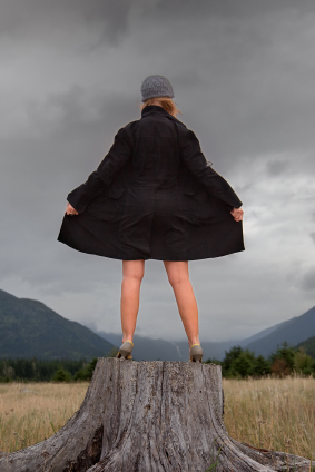 Woman in a trenchcoat standing on top of a tree stump and exposing herself to the world