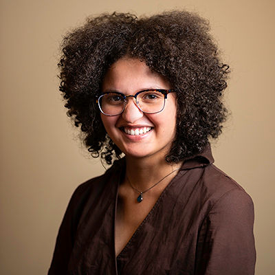 Image of a biracial Black woman with curly hair. She wears a brown top, a blue pendant necklace, and glasses.